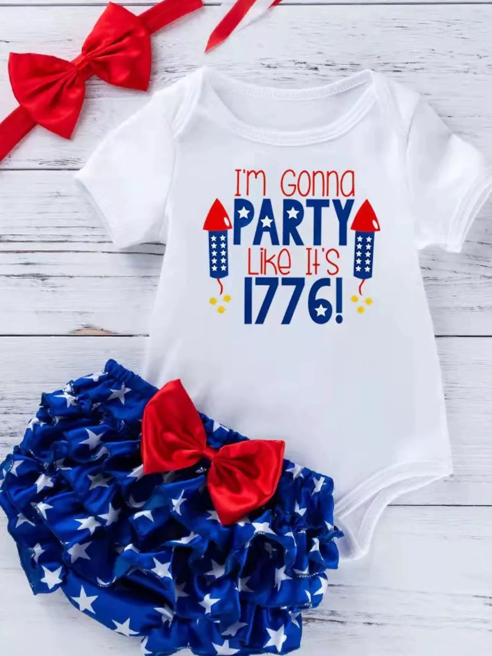 White patriotic baby onesie with red and blue text and ruffled bloomer with stars and red bow.